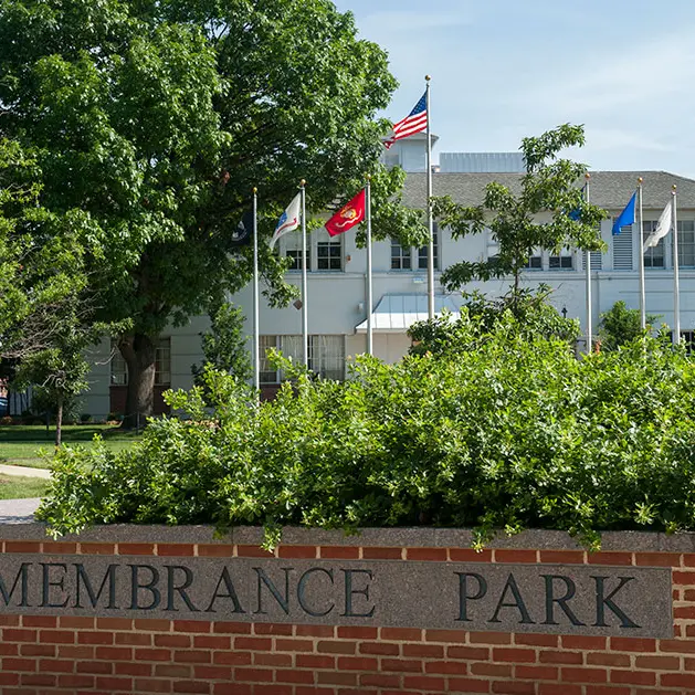 A brick wall that reads "Remembrance Park" sits in front of a large bush. There are flag poles in front of a white building. 