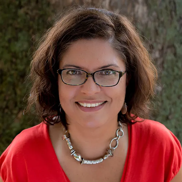 This woman poses for a photo with a pretty smile, glasses and a cool silver necklace. She looks both friendly and like someone who'd make a great friend.
