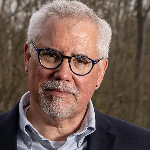 A man whose soft smile is reflected in his lips and eyes poses for a photo in the woods. He wears round glasses and a button-up under a suit coat, whose color is subtly reflected in the frames of his glasses