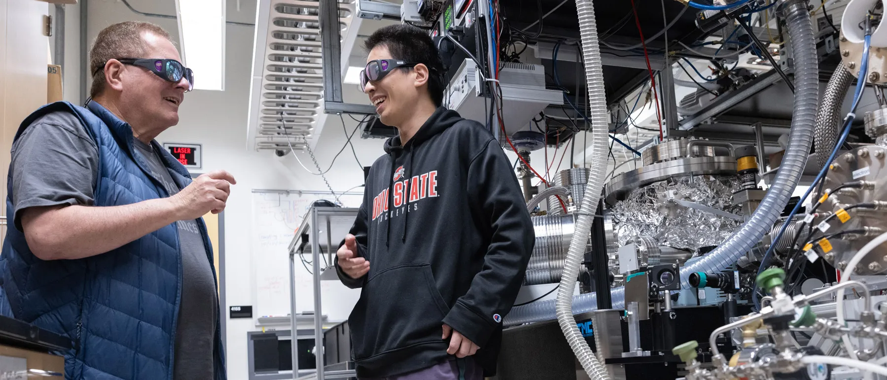  Lou DiMauro and Yaguo Tang talk in front of the Quantum Trajectory Selector Yagou works with. Plastic tubes swarm in front of the metal portion, which is several metal-cylinder-style components connected by smaller metal tubes. 