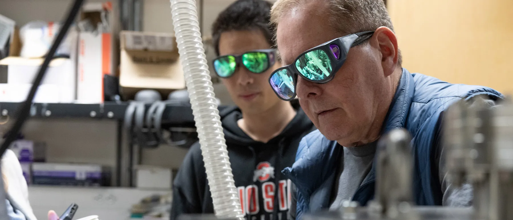 Both wearing safety goggles, Lou DiMauro and Yaguo Tang talk about a Quantum Trajectory Selector, which is the apparatus that lets them shoot lasers into gas to track electrons. Metal canisters and plastic tubes can be seen.