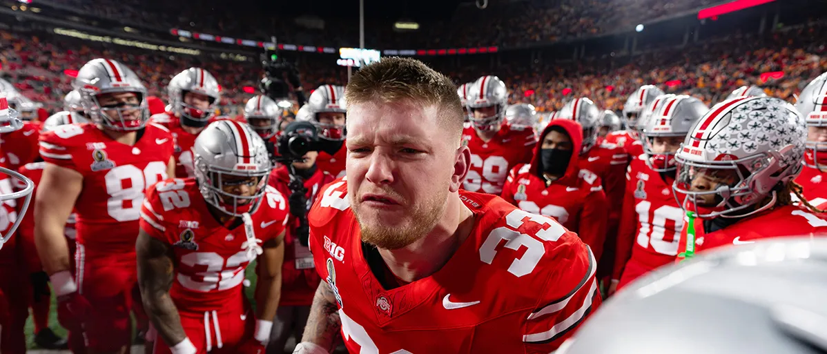 A young white man wearing hsi uniform but no helmet stands in the middle of a huddle of fellow players emotionally saying something. His eyebrows are furrowed and his short hair and beard are neatly trimmed