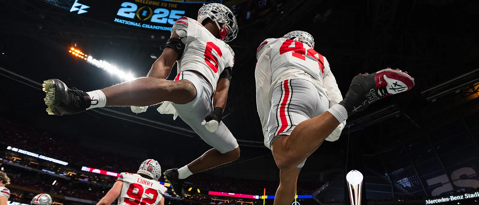 Taken from below as 2 football players launch into the air, this photo shows two young black men who seem to be casually flying around in midair. The sky above them is black, interrupted only by stadium lights