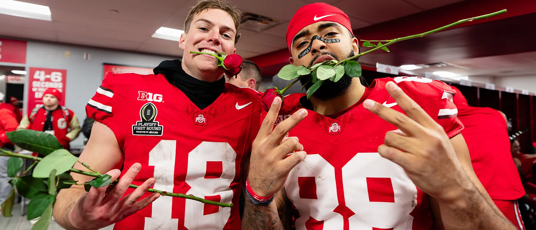 Two uniformed football players, one white, one black, both attractive young men, clench a rose each between their teeth and flash finger peace signs for the photographer