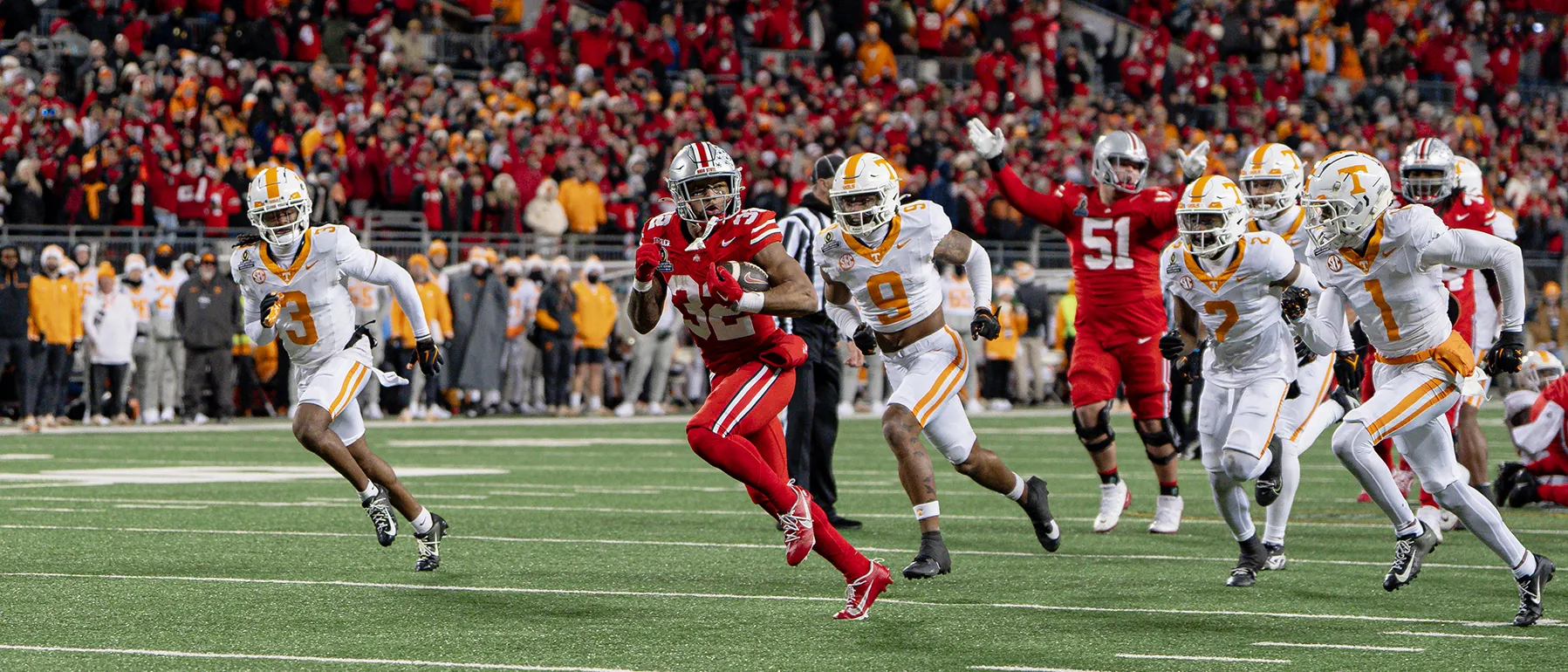 An Ohio State running back carries the football as five members of the other team give chase. The Buckeye is a young Black man with muscular arms and the type of powerful stride that appears effortless
