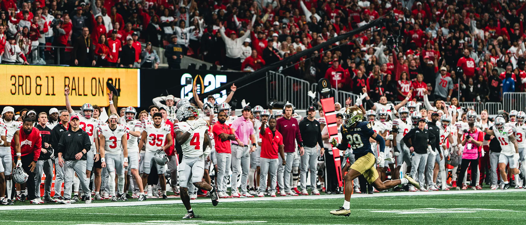 A sprinting Ohio State player looks back over his shoulder, preparing to catch a football thrown so far and so high, it's not even seen in the photo. The Buckeye is a young black man, his trailing opponent seems at least 10 yards behind him. On the sidelines behind them, the Ohio State team watches with anticipation, wide open mouths or dawning smiles