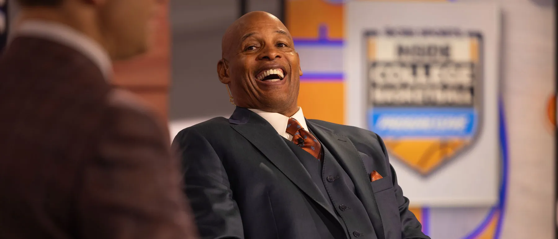 Kellogg laughs during an on camera interview in a television studio, with another person visible in the foreground and a sports themed backdrop behind them