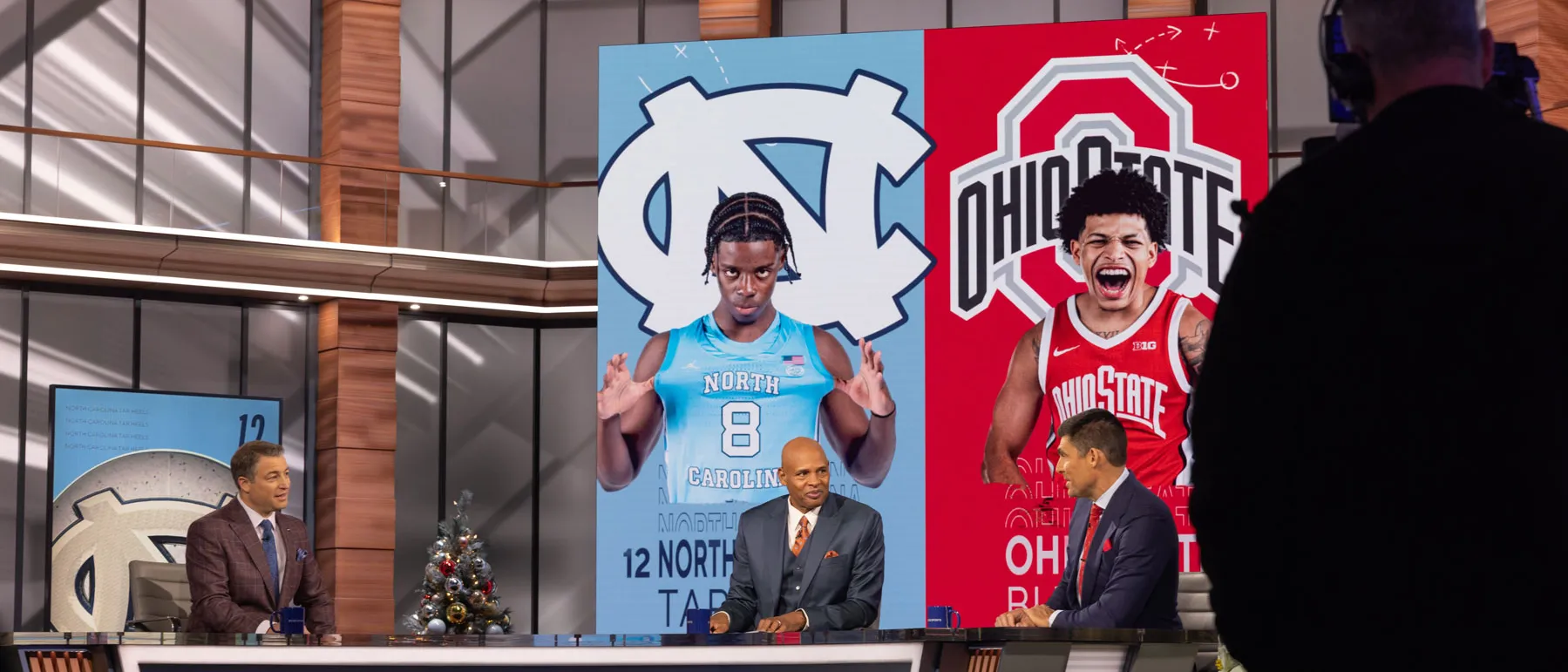 Three sports commentators, including Clark Kellogg, sit behind a studio desk during a televised sports segment, with large graphics of two basketball players displayed on screens behind them. One is Ohio State player John Mobley Jr. A camera operator stands in the foreground, and holiday decorations are visible around the set.