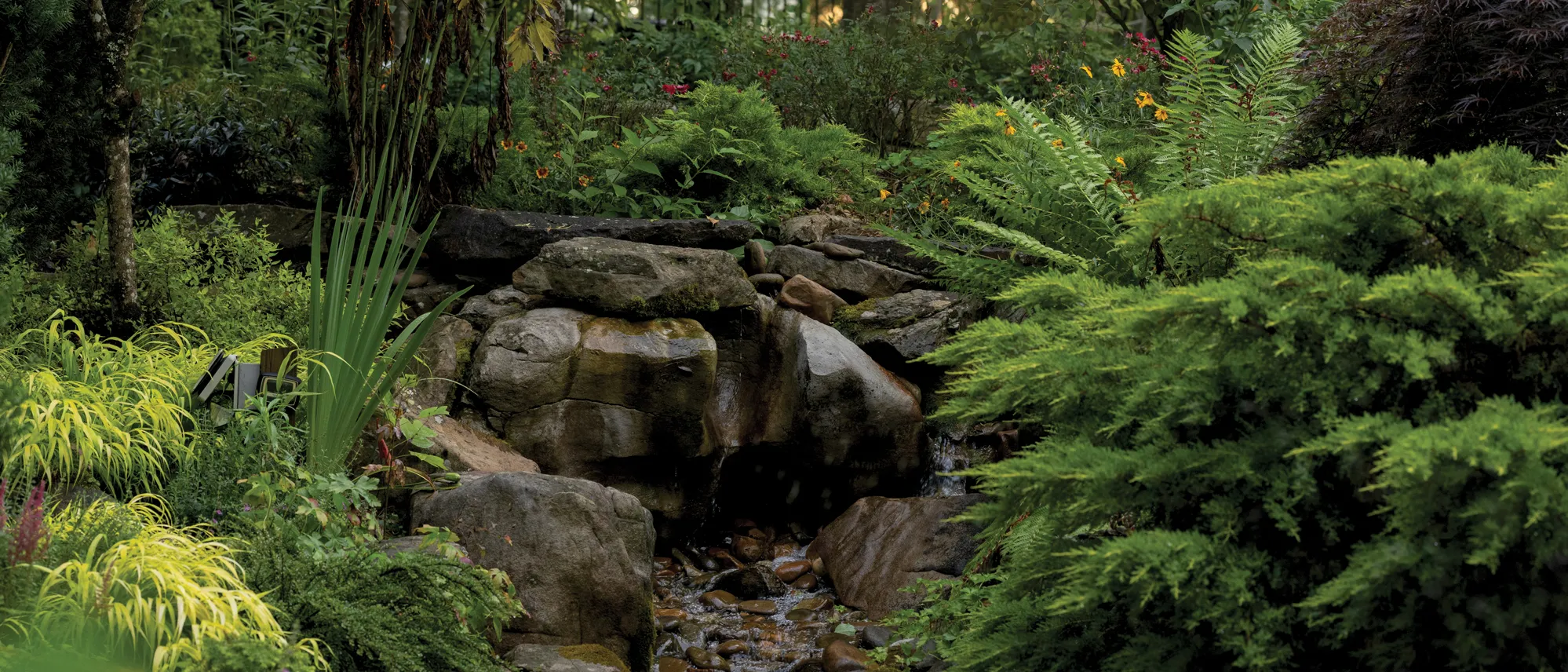 An artful rock formation spouts a bubbling stream. Flowers, ferns and other greenery surround it.