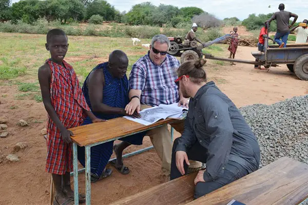 A group of people are sitting at a small wooden table looking at a large piece of paper. 