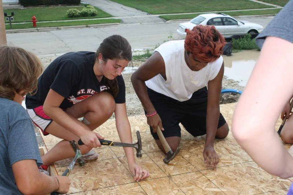 While a college student, Andre kneels to nail wood on what will be a porch floor. He has pink-dyed hair and shorts and a T-shirt. Students around him similarly work.
