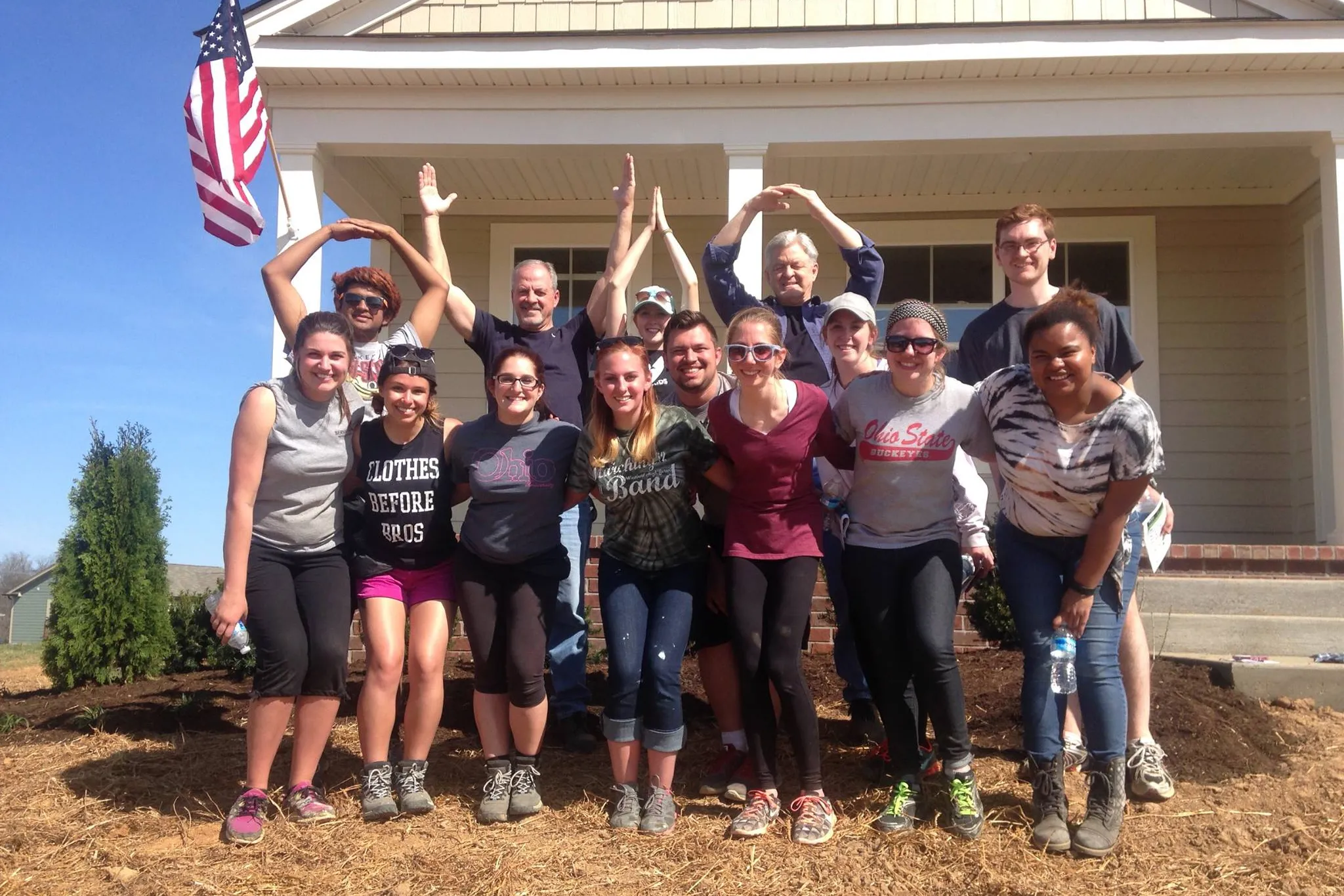 A group of students and teachers cheers in front of a completed Habitat for Humanity house. Andre stands in the back row making the O for the O-H-I-O.