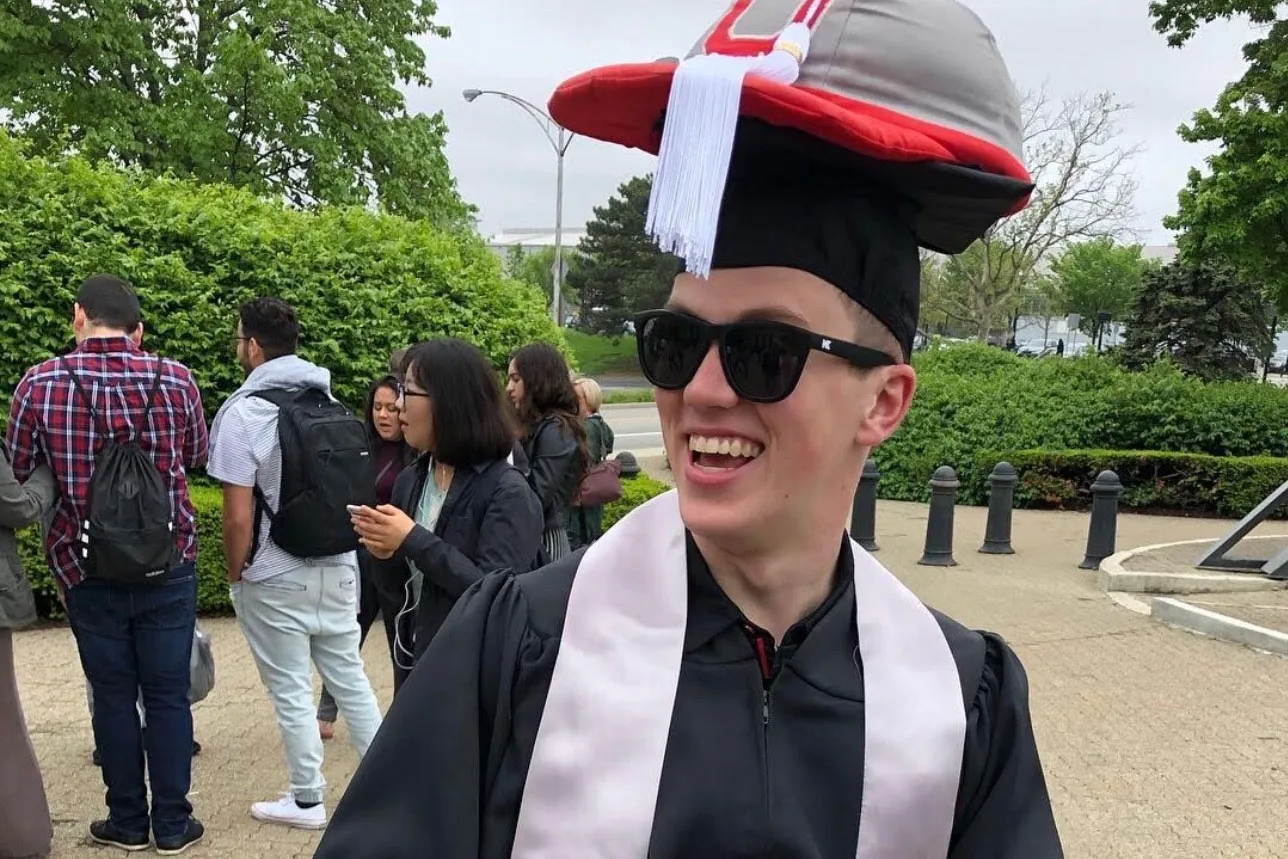 A graduate wears Brutus hat on top of his graduation cap and sunglasses. He seems cool and happy.