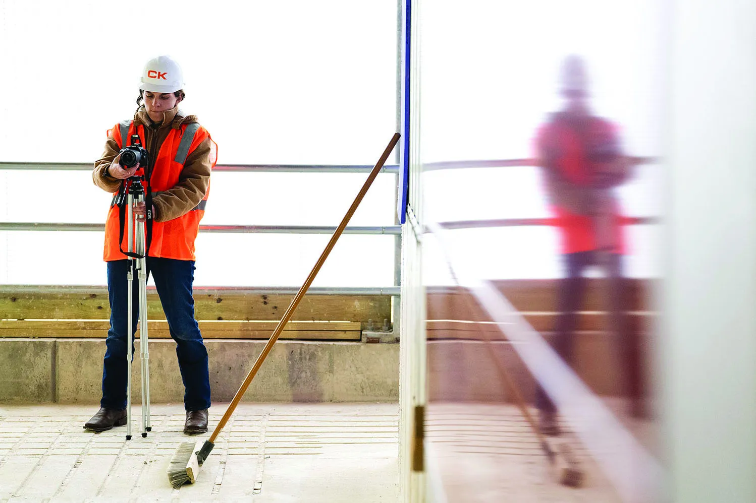 Wearing a safety vest and hard hat, a young woman focuses a camera on a closed tripod within a construction site.