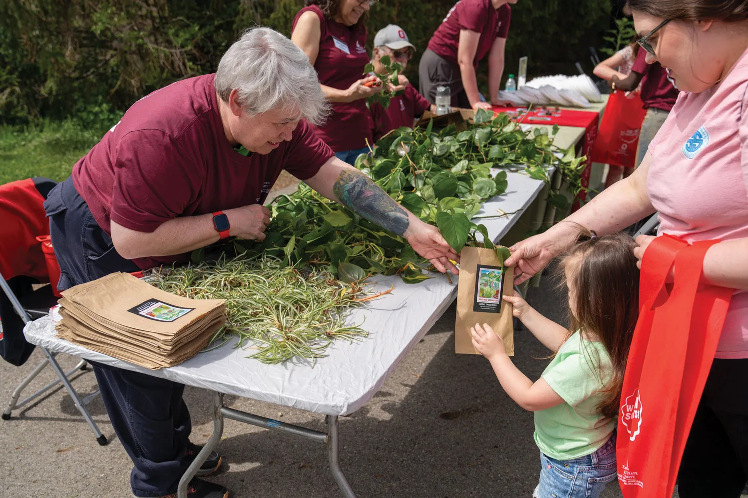 Leaning over a folding table covered in plant cuttings, a woman hands a brown paper bag to a young child, who's supported by her mom standing behind her. They're all smiling.