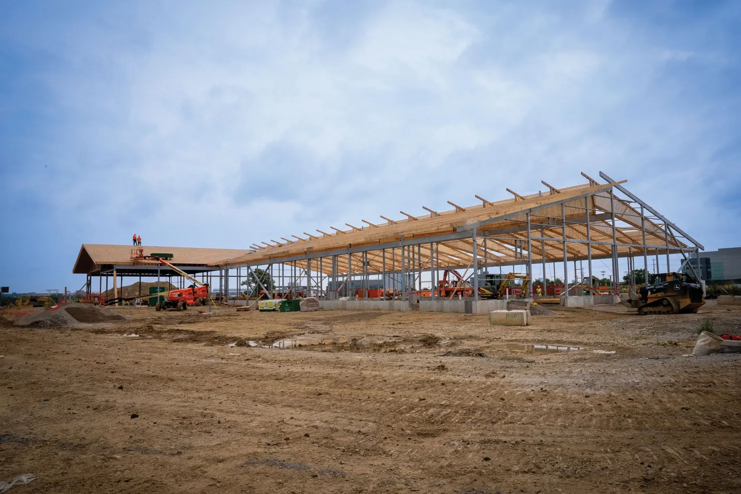 Against a pretty blue sky, framework for a new long but low building rises. A yellow tarp hangs over the roofing while the vertical metal frame is bare, creating a striking silhouette. 