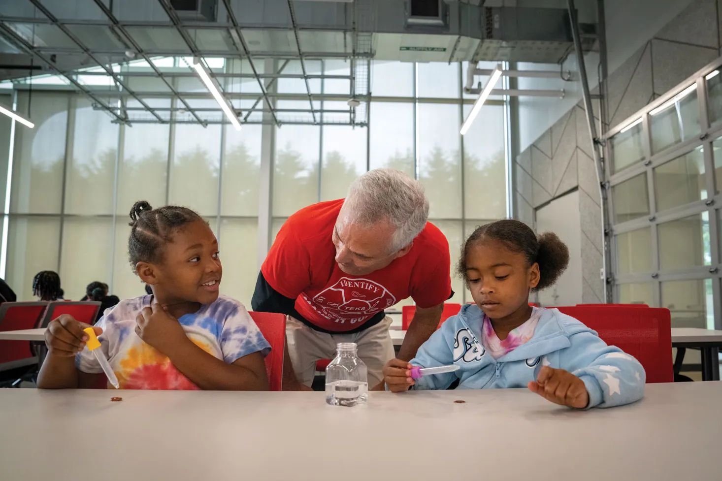 Backed by a wall of windows and tables full of kids, two young Black children sit at a table with small instruments. A white man leans between them and laughs with the girl he's speaking with.