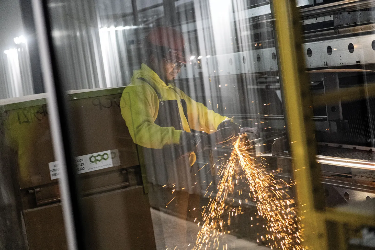 Behind a window and along side techy-looking equipment, a worker wearing a safety vest 
