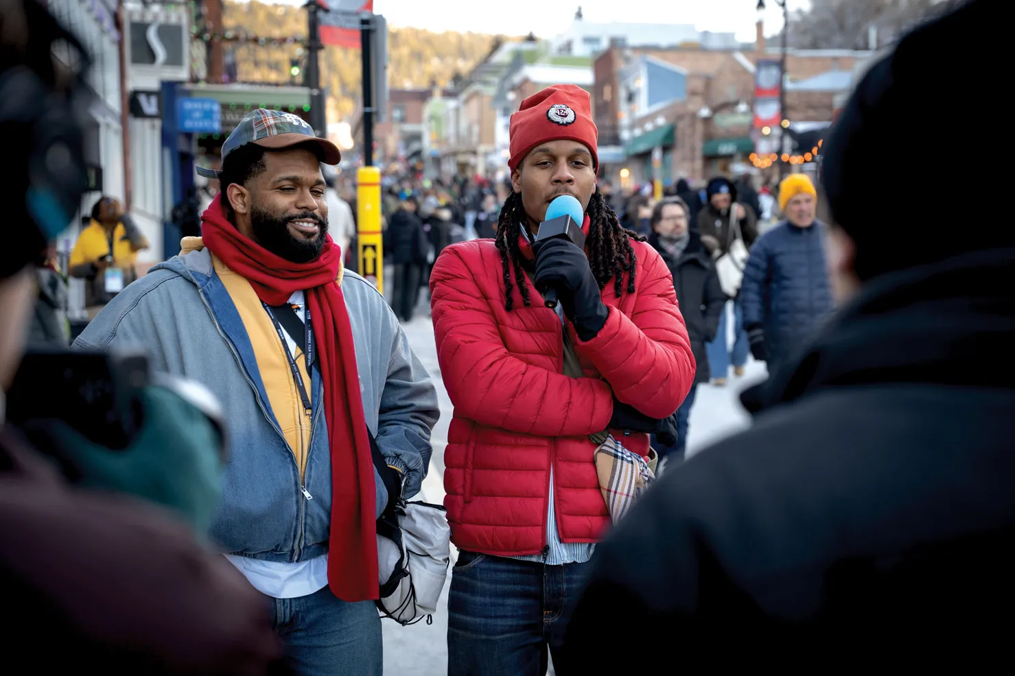 Justen holds a mic close to his mouth as he speaks to Tv interviewers in front of him--only their backs can be seen in this photo. His brother Julien smiles, seemingly in agreement. They both wear coats and hats and the street scene behind them looks both quaint and cold.