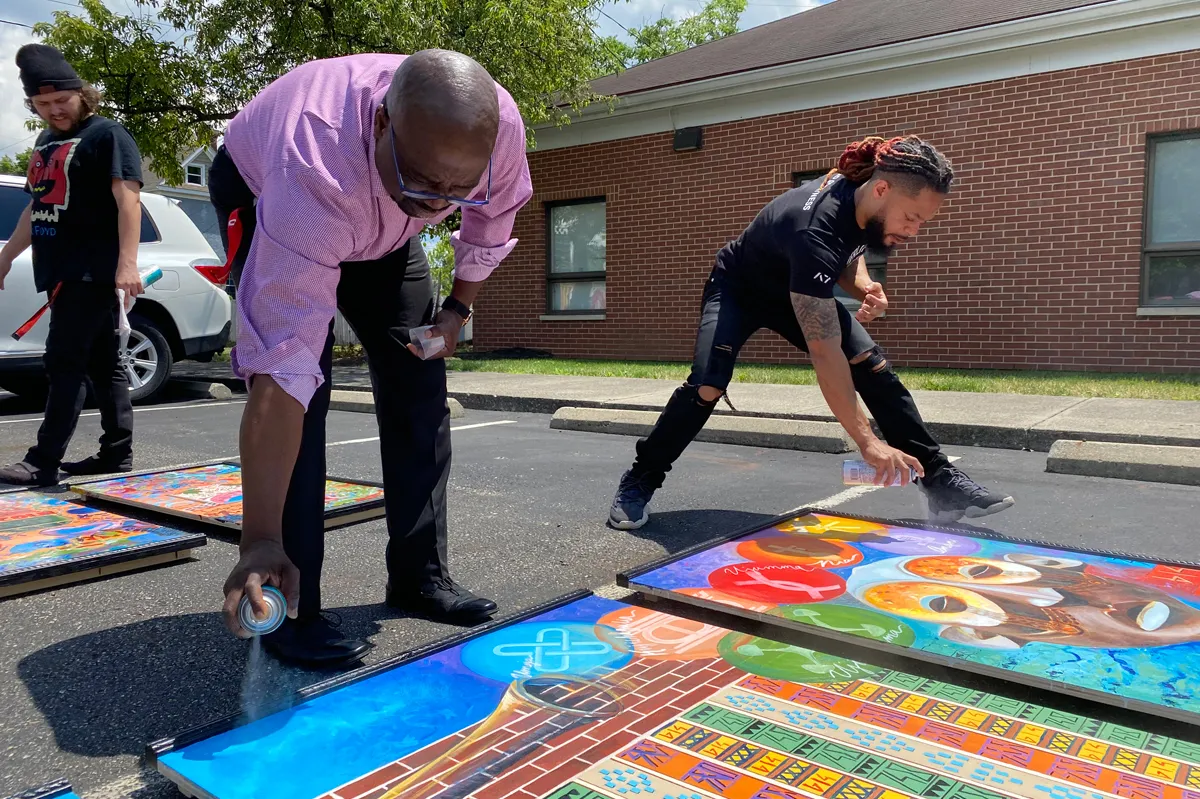 Men use spray paint to create large scale artworks lying in a parking lot. Terron Banner is at right.