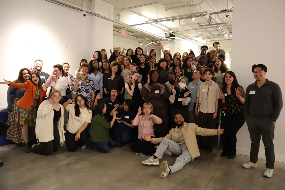 50 or so people pose for a photo. Banner is sitting on the floor at front right and has his arms spread wide