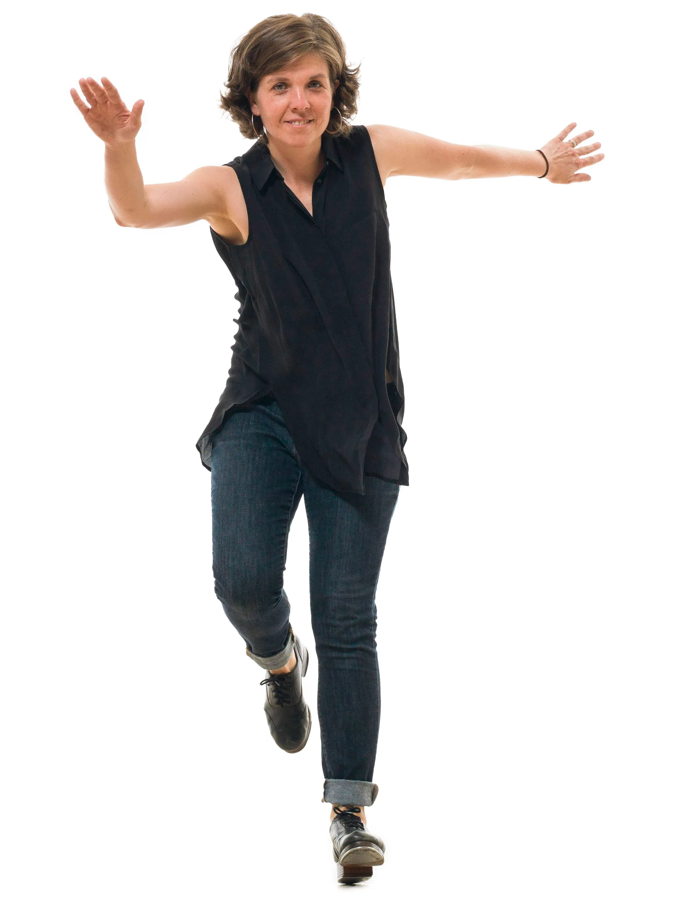 Jenai Cutcher, a white woman with chin-length hair, smiles as she dances in her tap shoes. Her hands are stretched wide apart and one leg is bent to the degree that that foot is off the ground.