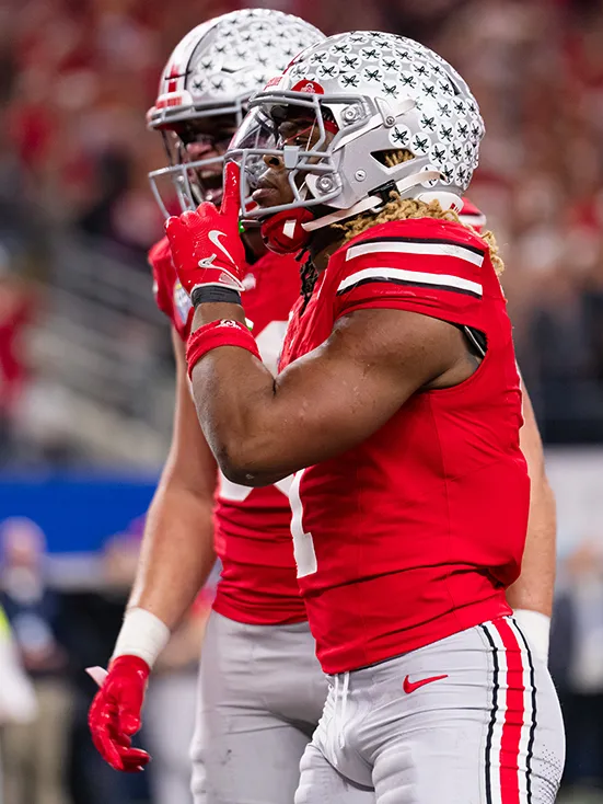 A pair of Buckeyes are on the field with their helmets on during a pause in play. The one closer to the camera can be seen better. He is a black man with shoulder length braids that are extra fun. The ones closer to the camera are blond, while those on his other side are black.