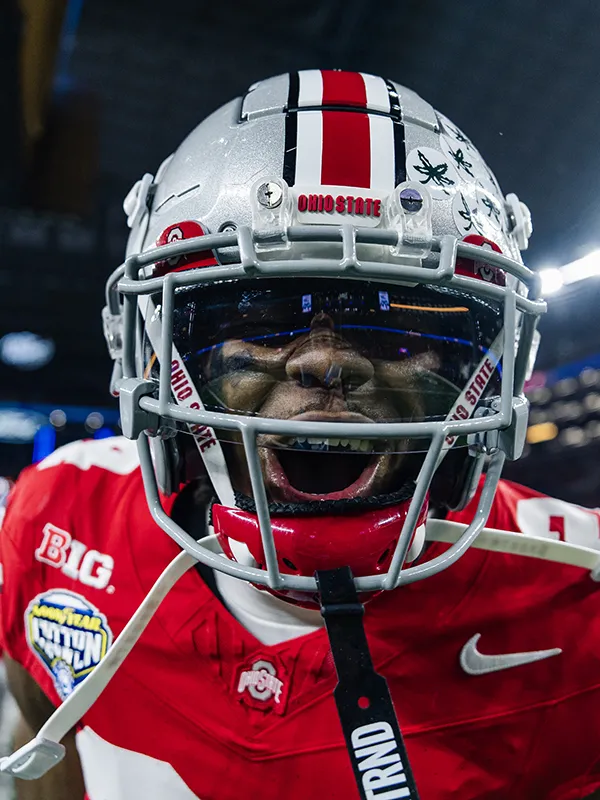 A young black man wearing his football helmet happily calls to the photographer. The photo is a close-up of his face, though his eyes can't be seen well because of lights reflecting in the protective see-through screen covering the top half of his face.