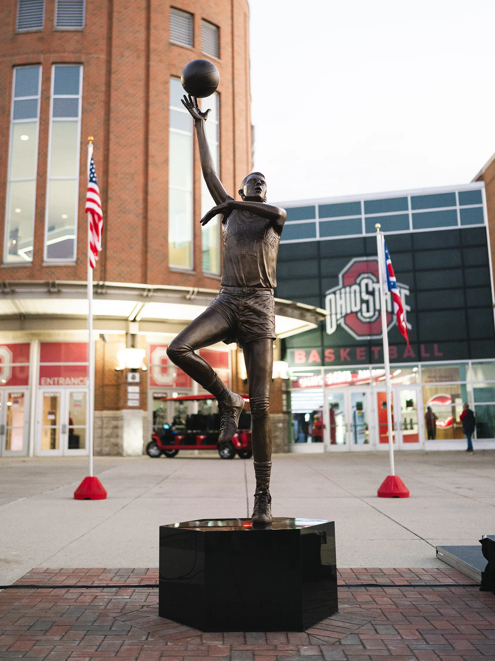 The statue of Jerry Lucas outside the Schottenstein Center is bronze and bigger than life size. Jerry lifts his right leg, extending his left with his weight there. His right arm is almost fully extended but slightly rounded as he rolls the basketball off his fingertips. His left arm is bent loosely in front of his torso. His open-mouthed expression shows his focus.