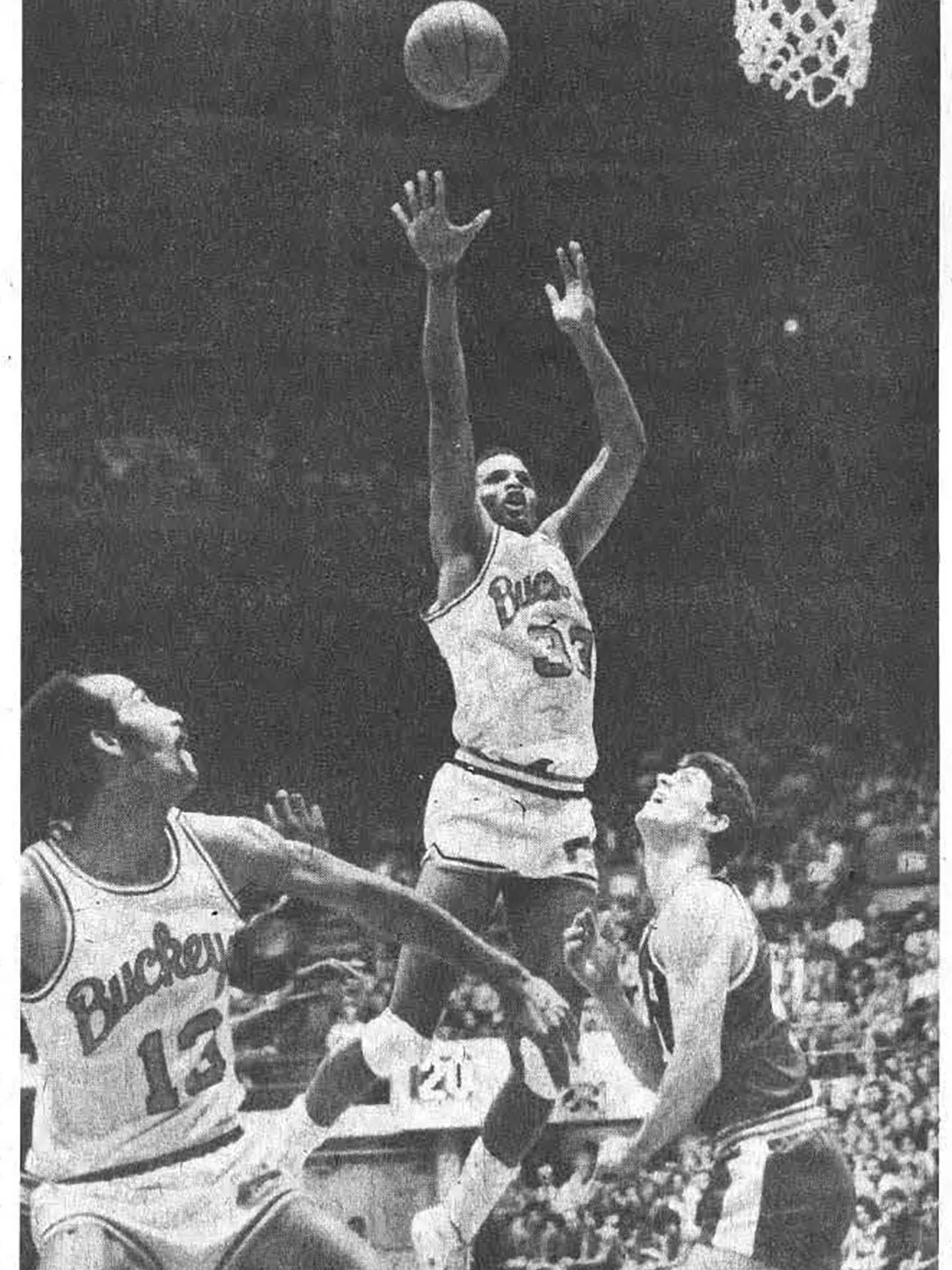 As an Ohio State player, Kellogg wears a Buckeyes uniform and jumps high over other players, shooting at the basket with arms extended. Two other players stand nearby on the court, one in a Buckeyes uniform and one in another team’s uniform, watching the play under the hoop.