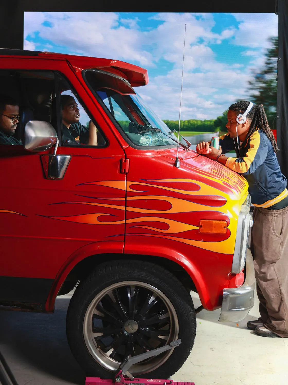 Justen stands in front of a van slightly leaning over its hood to talk with actors in the front seats. He wears a headset and meets their eyes. The van is scarlet with yellow flames.