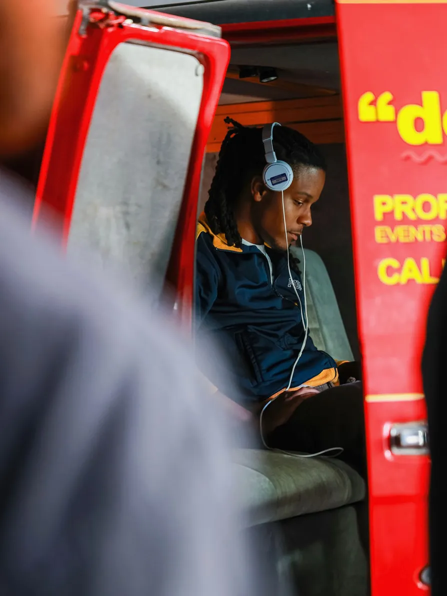 Justen works on a computer while sitting in the backseat of a van that is part of the scene that is filming. He has headphones and intense focus.
