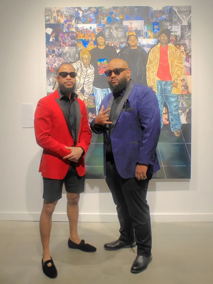 Banner poses with another man in front of a painting of Black men amid a street scene. Both are dressed in striking tuxedo-style coats and wear sunglasses.