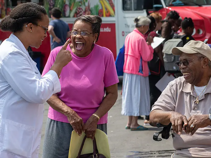 Dr. Sophia Tolliver is wearing a white lab coat while speaking to 2 of her patients. The patients are a woman in a pink shirt and a man in a tan ball cap and shirt who is seated. 