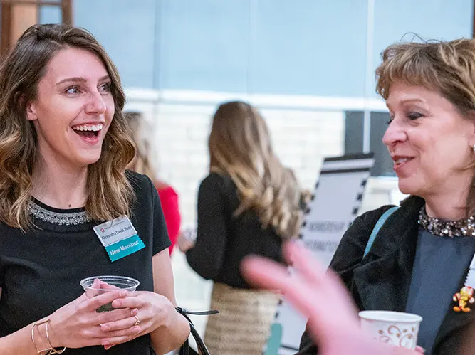 Two women are in a discussion while at an event. They are wearing names tags and holding disposable cups., More than 200 women comprise the membership of Women &amp;amp; Philanthropy, a giving circle-style philanthropic organization dedicated to funding researc