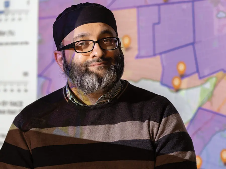 A bearded man wearing glasses and a kufi looks directly at the viewer and slightly smiles. A colorful map is projected onto him and the wall behind him. 