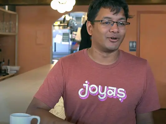 An American man of Indian descent smiles while chatting as he leans on the counter in his restaurant. He looks like he's enjoying himself.