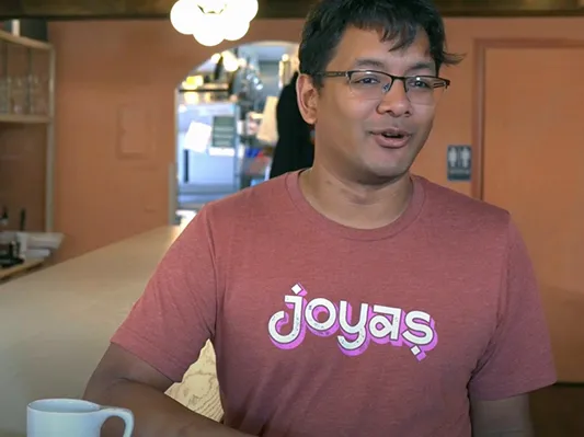 An American man of Indian descent smiles while chatting as he leans on the counter in his restaurant. He looks like he's enjoying himself.