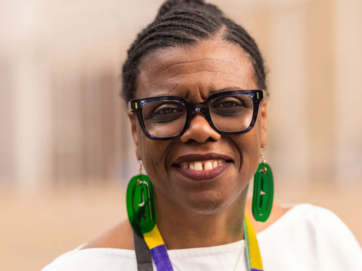 Gaëtane Verna, a Black woman with her braided hair pulled back in a bun, is shown from the shoulders up, smiling as she seems to make eye contact with the viewer outside of the Wexner Center for the Arts. She wears thick-rimmed glasses and big transparent earrings, adding to her confident, artsy presence.