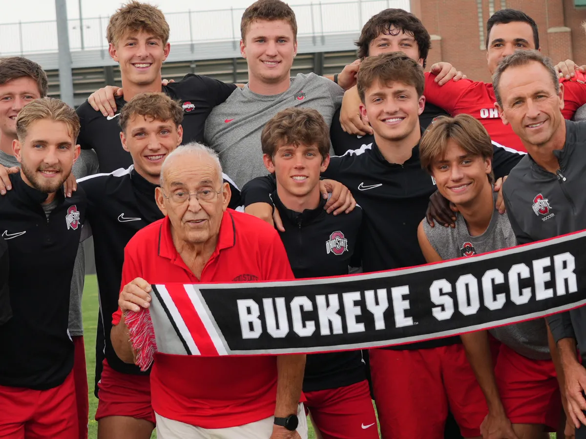 Paul Halpern, a 91-year-old man, poses with Ohio State's men's football team and coach on the soccer field at Schumaker Complex. They hold a banner that reads Buckeye soccer