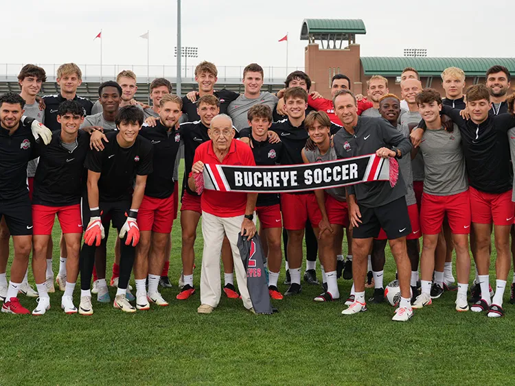 Paul Halpern, a 91-year-old man, poses with Ohio State's men's football team and coach on the soccer field at Schumaker Complex. They hold a banner that reads Buckeye soccer