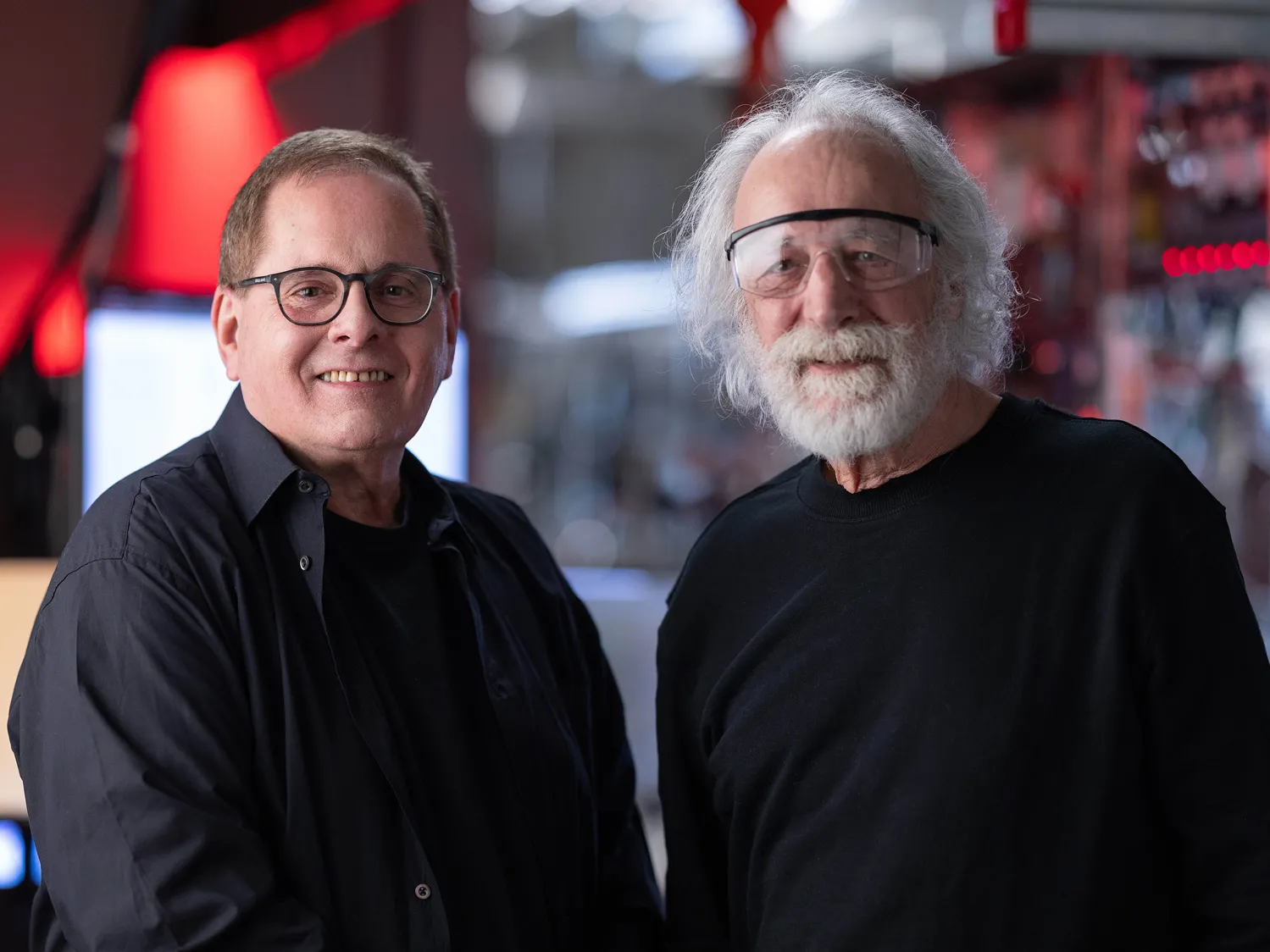 Lou DiMauro and Pierre Agostini smile as they pose for a portrait. Pierre has flowing white hair and a beard and wears safety goggles. Lou has short brown hair and glasses. Both are white men who radiate friendliness while dressed casually. 