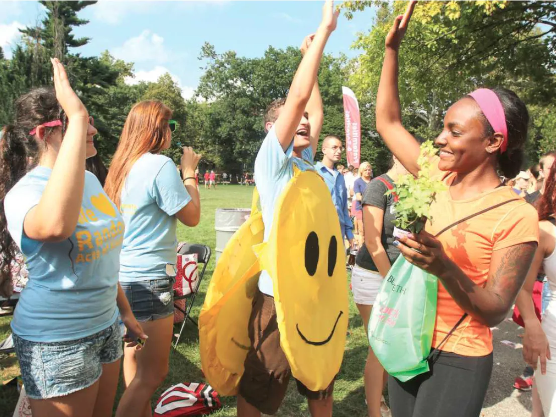 Ohio State students on the Oval give high fives. The sun shines brightly and most wear summer clothes. The guy in the center, though, wears giant smiley faces on his front and back.