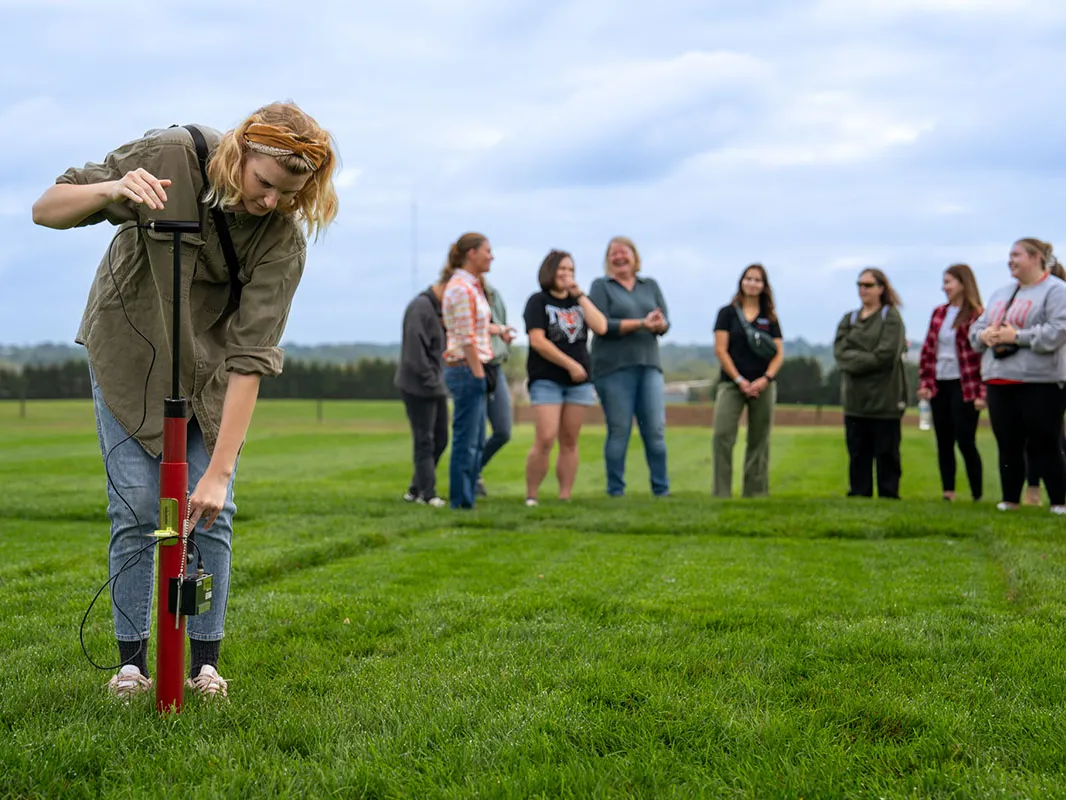 a student uses a turf tool that looks vaguely similar to a pogo sticks. In teh distance a line of students stands and watches