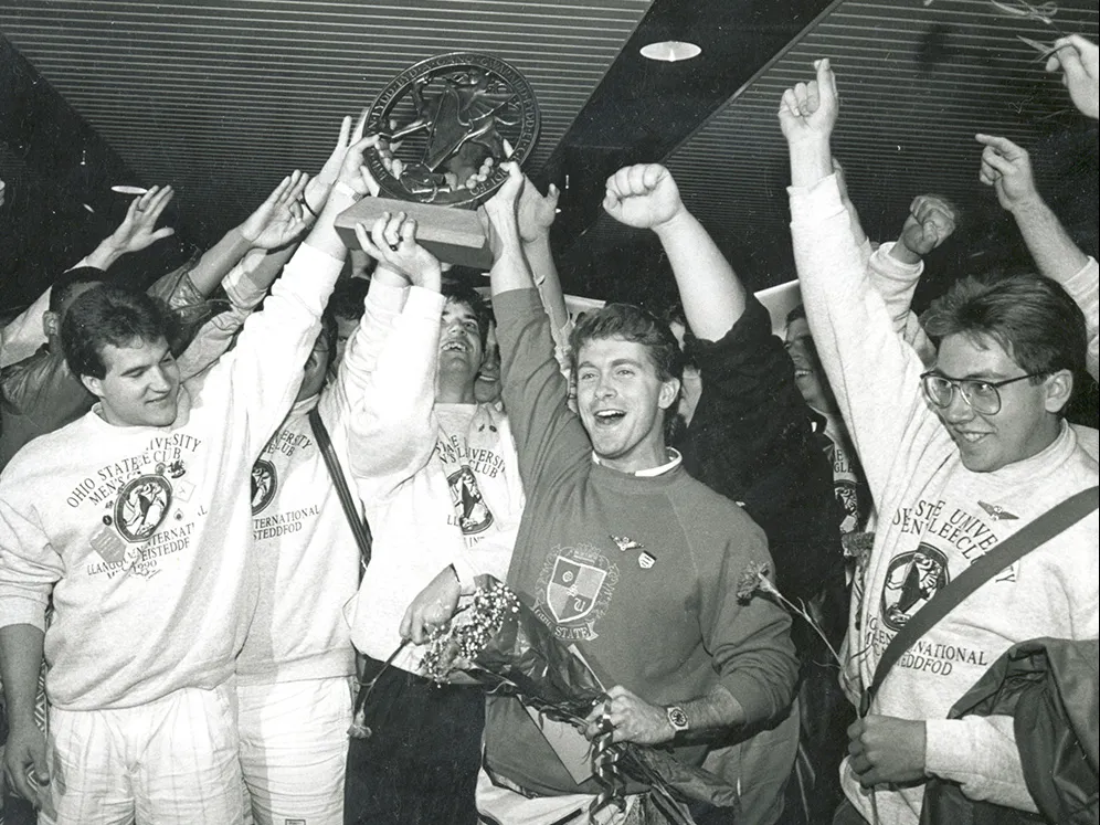About a dozen members of the men's glee club hoist a trophy as they cheer. The photo is black and white, giving it a dtaed look, even though color photography was available at the time.