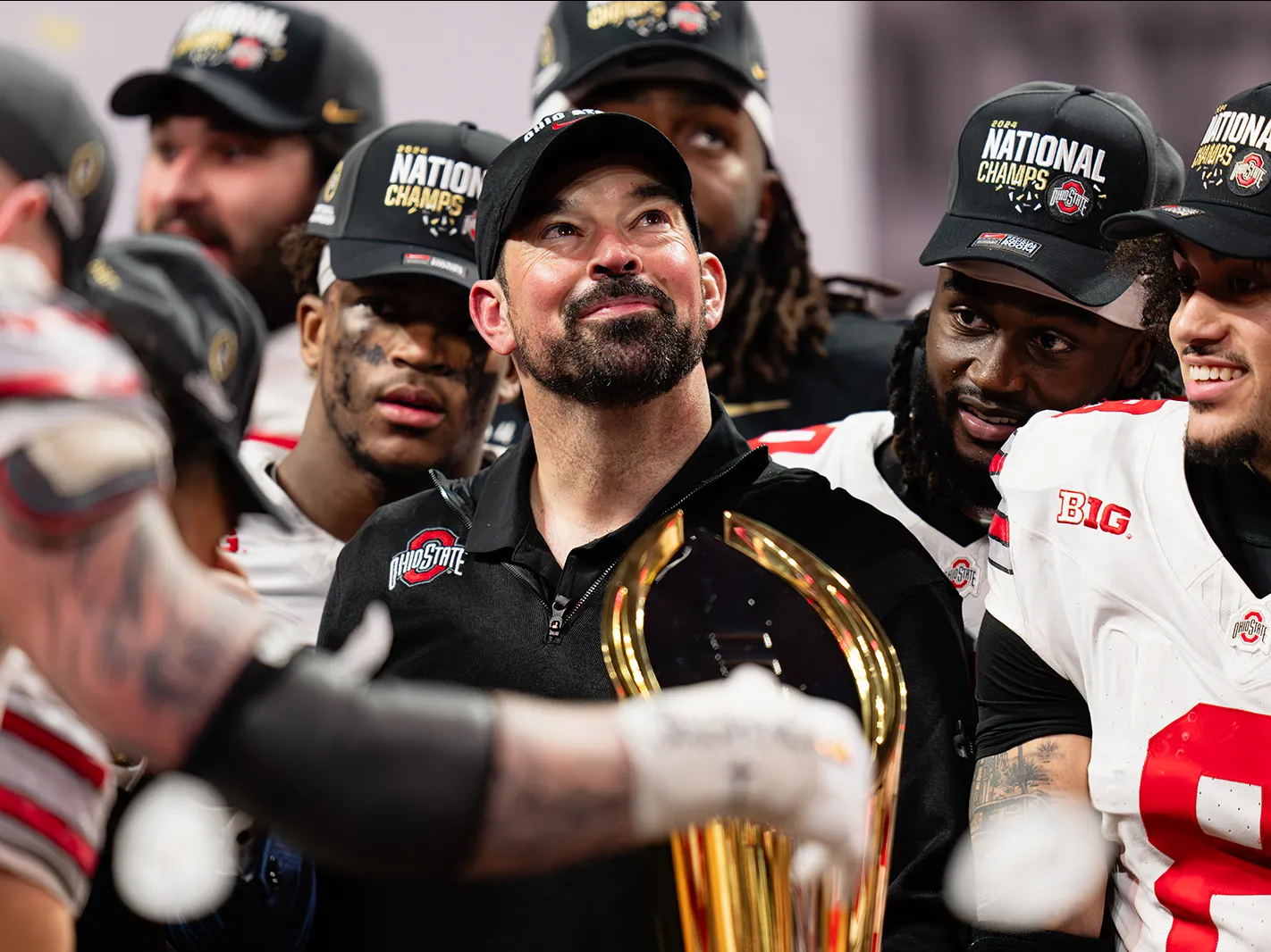Ohio State football coach Ryan Day, surrounded by football players, looks proudly upward. He appears a little tearful but is smiling. Everyone in the photo taken after they won their final game of the playoffs wears a National Champs baseball cap.