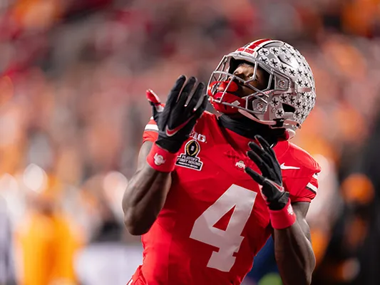 A young Black man wearing a football uniform looks back over his shoulder as he runs forward. His hands are cupped and ready to catch teh football and his helmet is coated in Buckeye stickers, which were each awarded for a fabulous play in a game this season
