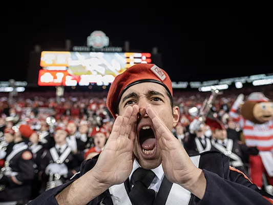 A young white man wearing a band uniform cups his hands around his mouth as he shouts. Dozens of band members and Brutus Buckeye can be seen behind him
