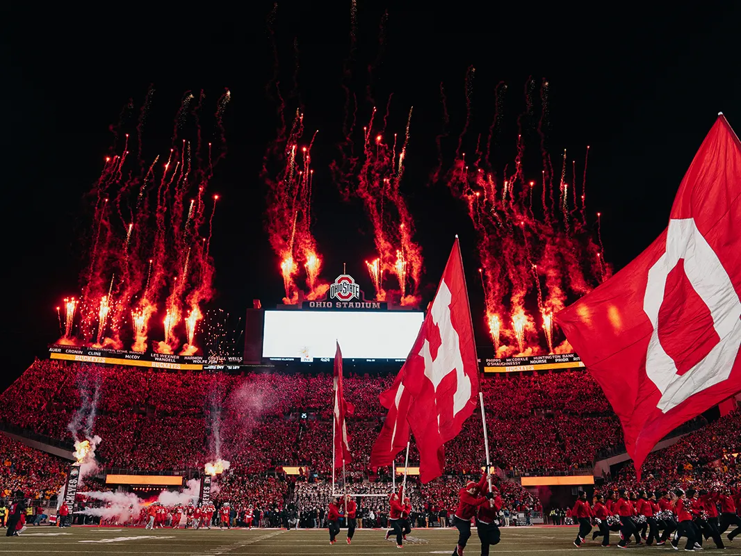 A photo taken from the field inside Ohio Stadium on a dark night shows the stands full of red-clad people who appear as dots and people running on the field with giants flags that spell out OHIO. Fireworks shoot into the black sky.