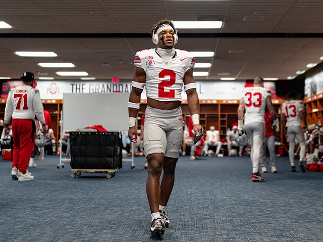 Inside a nice locker room, a Buckeye player, a young Black man wearing headphones that cover his ears and a warm-weather version of the the team's uniform, strides toward the camera but looks off to the side. He seems very focused on getting focused.